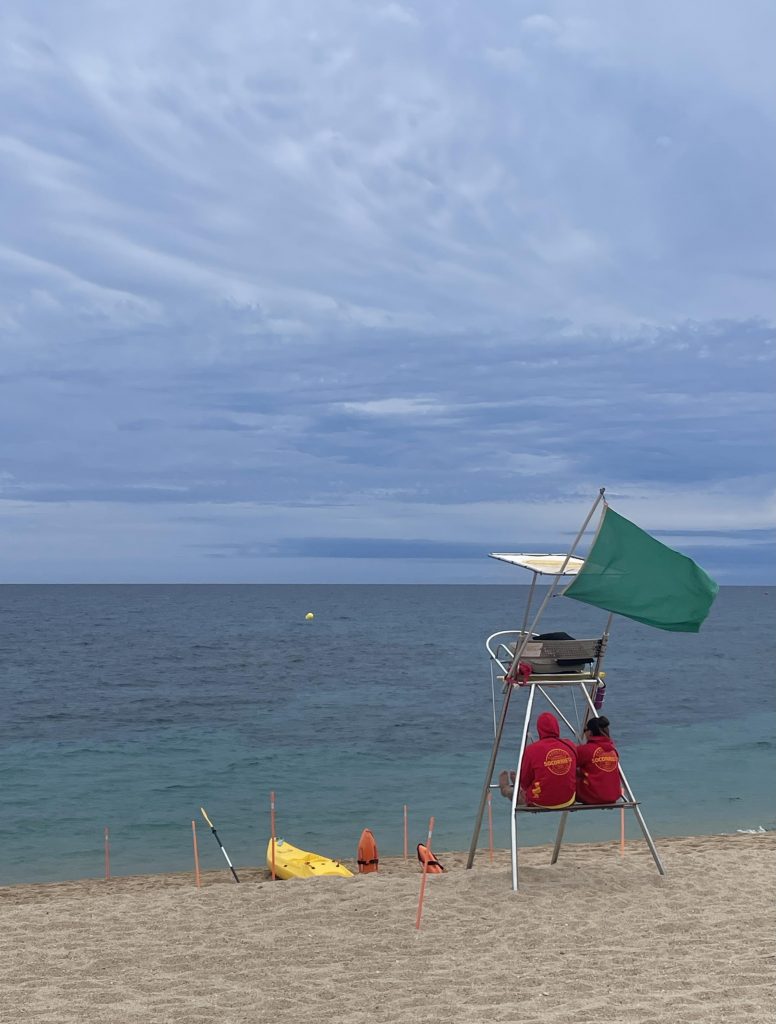 Strandszene bei Tageslicht, aber ohne Sonnenschein. Der Himmel erscheint zwar blau, ist aber mit Schlierenwolken überzogen. Dunkelblaugrau im Vordergrund Sandstrand. Auf diesem Strand steht ein Gestell, auf dem etwas erhöht zwei Personen in roten Sweatshirts mit Kapuzen sitzen. Eine Person hat die Kapuze über den Kopf gezogen. Die Person rechts neben ihr hat dunkle Haare, die zu einem Zopf geflochten sind. Das Gestell hat auch eine Überdachung. Vor dem Gestell sind orange Rettungsbojen in den Sand gesteckt. Und ein weißes Bord liegt auf dem Sand. Am Gestell flattert oben eine grüne Flagge. Weitere Personen sind am Strand oder im Wasser nicht zu sehen.