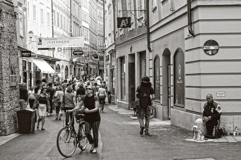 Eine Szene aus einer Fußgängerzone, in Grautönen. Es sind einige Menschen unterwegs, ein Fahrrad wird geschoben. Es hängen Werbeschilder über der Straße.