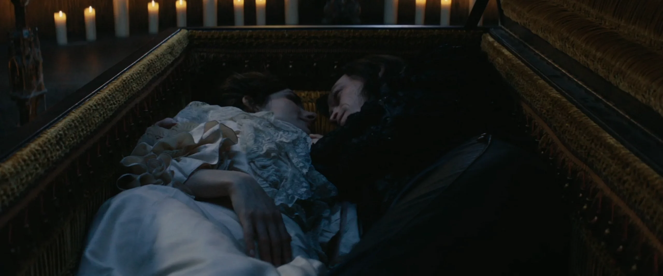 A man and a woman in period clothes lying together comfortably in a big coffin, facing each other