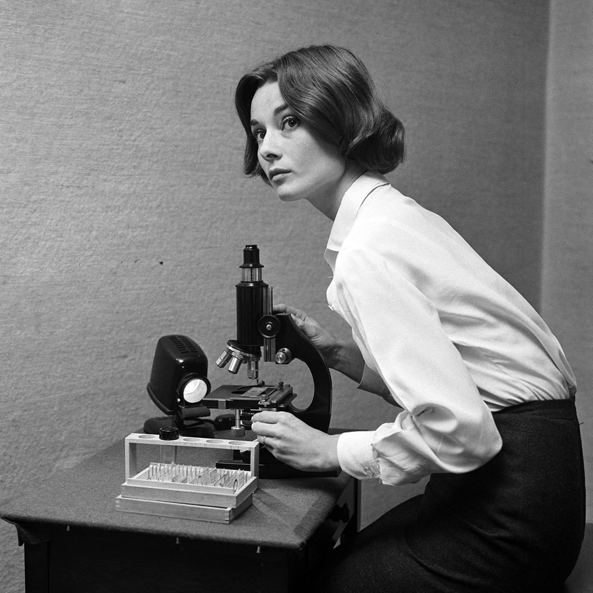 Black and white image of Audrey Hepburn sitting at a microscope