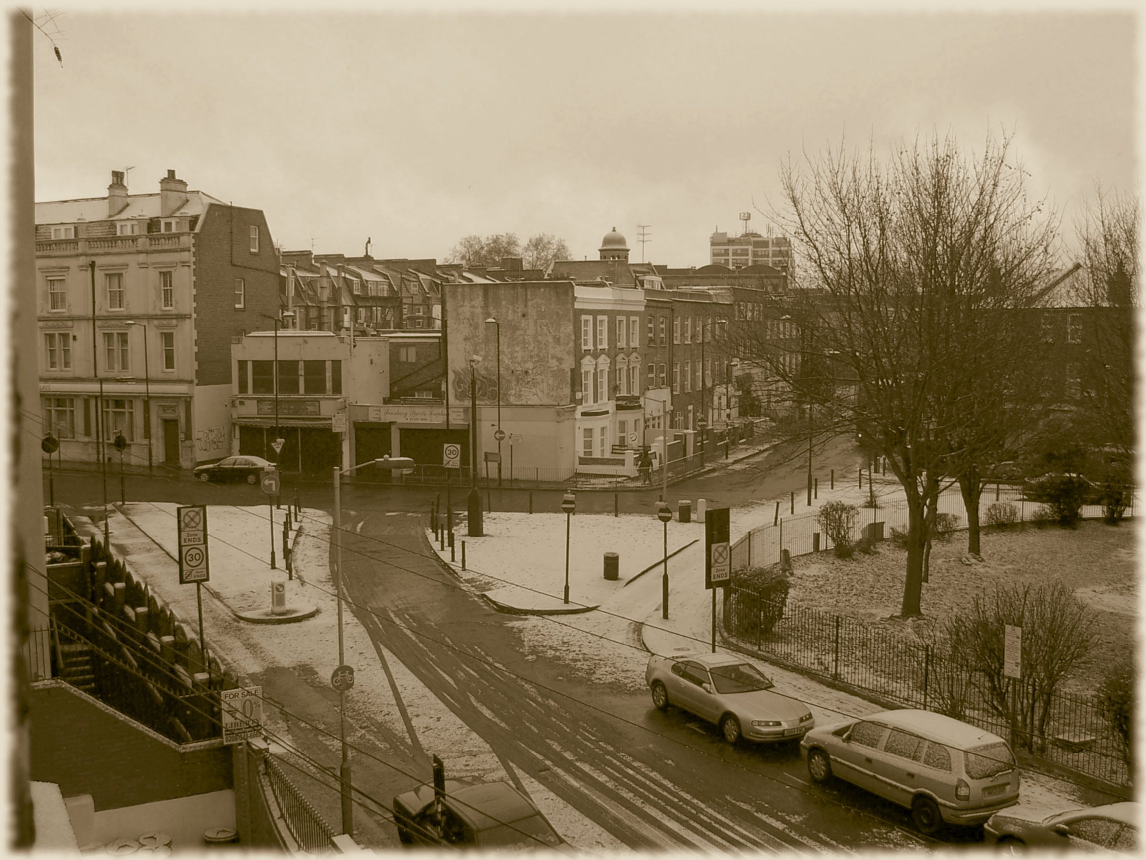 Ein Schwarz-Weiß Bild auf eine Straßenecke im Winter, rechts ein kleiner Park mit kahlen Bäumen in der Mitte 3-stöckige Gebäude aus dem 19., und ein Flachbau aus dem 20. Jahrhundert. Auf allem liegt eine dünne Schneeschicht.