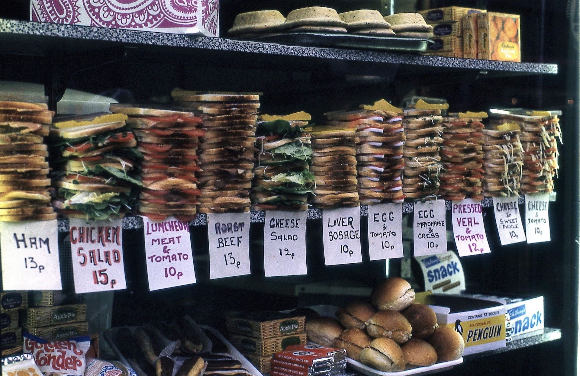 Food for sale in London 1972 on three shelves. The remarkable part is that on the middle shelf, there are piles of unwrapped sandwiches, each pile labelled with a hand written sign.