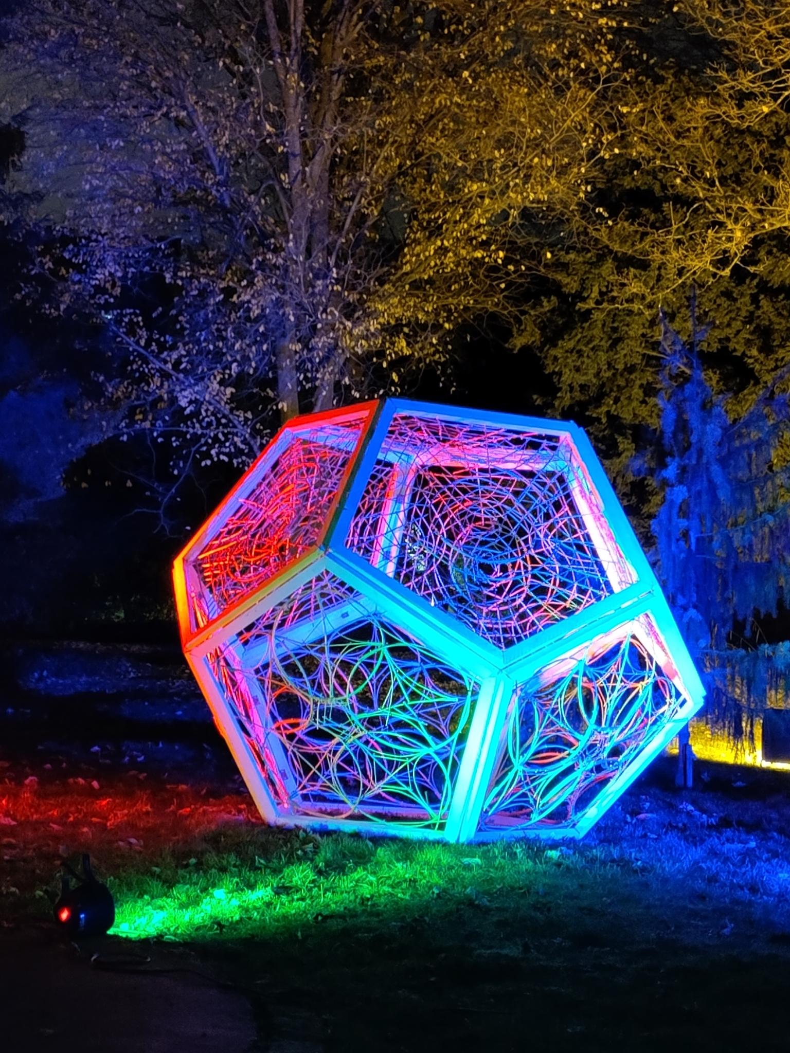 Eine Kunstinstallation in einem Park, im Hintergrund sind Bäume. Das Objekt hat 12 Seiten die jeweils 5 Ecken haben. Die Flächen sind zusätzlich mit Mustern verziert. Das Installation wird von verschiedenen Seiten bunt angestrahlt.