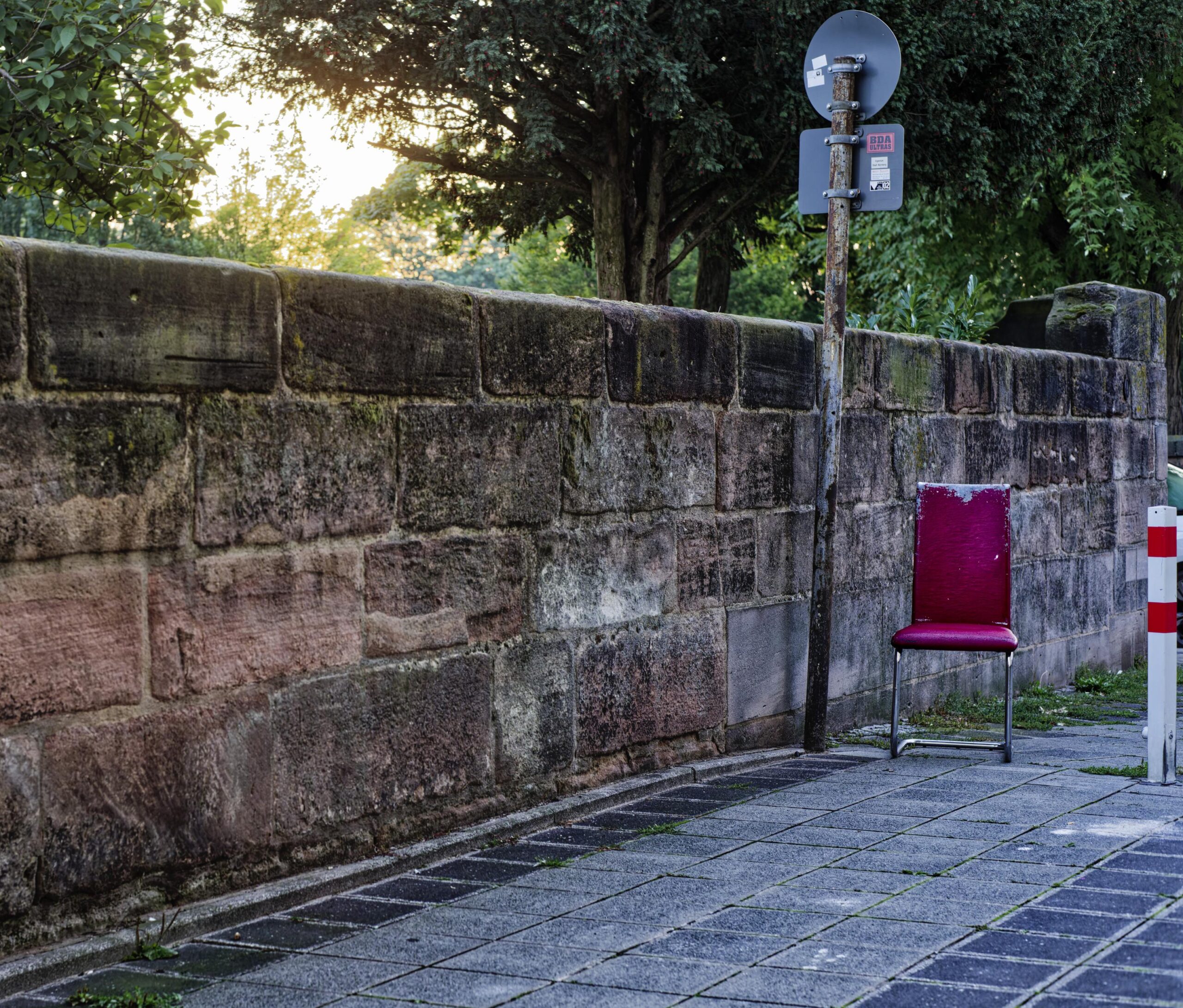Das größte Objekt ist eine Mauer aus dem in Nürnberg typischen Burgsandsstein. Davor geht perspektivisch ein Fußweg mit schmuddeligem Pflaster nach rechts hinten ab. Man ahnt das vom Bildrand abgeschnittene Ende des Wegs; ein rot-weißer eckiger Pfosten verhindert das Durchfahren von Autos. Zwischen dem Pfosten und der Mauer steht ein abgenutzter, weinroter, gepolsterter Stuhl mit Stahlrohrgestell anstelle von Stuhlbeinen. Links daneben ein Straßenschild, das man von hinten sieht. Hinter der Mauer sind Bäume zu sehen, und man sieht das Licht der aufgehenden Sonne.