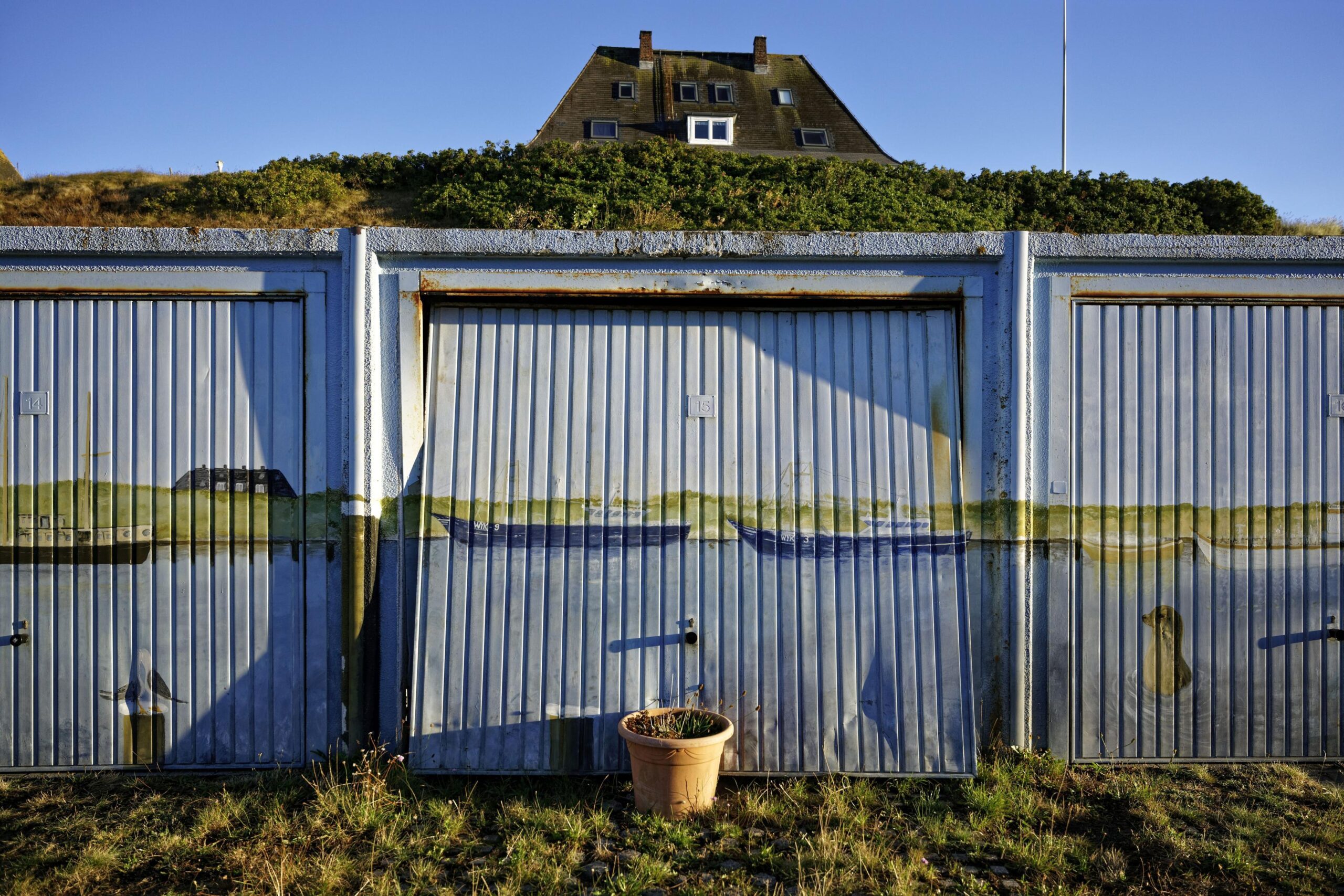 Hauptmotiv: Ein abgerocktes Garagentor, rechts und links schließt sich je ein weiteres, nur halb sichtbares, an. Alle sind bemalt mit einfachen Küstenmotiven (Schiffe, eine Robbe und ein Hausdach hinterm Deich). Vor dem mittleren Garagentor steht ein Blumentopf mit halb vertrocknetem Lavendel, der verhindert, dass das leicht geöffnete Tor weiter aufgehen kann. Vor den Garagen Gras, dahinter eine Düne mit Syltrosen, hinter der das Dach eines Hörnumer Kapitänshauses aufragt. Oben ein kleiner Streifen blauer Himmel.