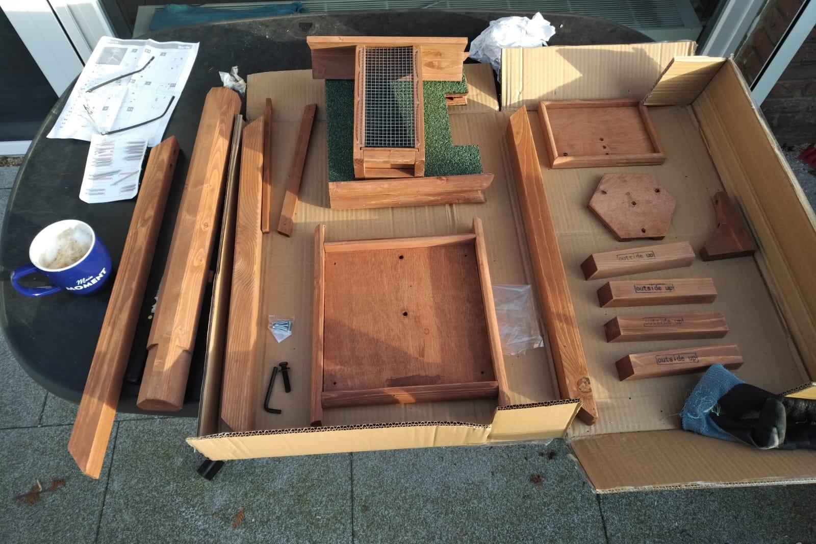 Auf einem Tisch liegt eine große geöffnete Kartonverpackung. Auf und neben dieser liegen geordnet zahlreiche vorwiegend hölzerne Teile eines Bausatzes in verschiedenen Größen und Formen. Außerdem ein Beutelchen Schrauben, ein Handschuh, ein kleiner Inbusschlüssel und eine große Schraube. Neben dieser Anordnung, die fast den kompletten Tisch einnimmt, finden sich am Rand des Tisches noch eine große blaue Kaffeetasse, frisch gefüllt mit Kaffee und Schaum, sowie eine unleserliche Aufbauanleitung, auf der eine Brille liegt. Der Boden unter dem Tisch und das Sonnenlicht zeigen, dass er draußen steht.