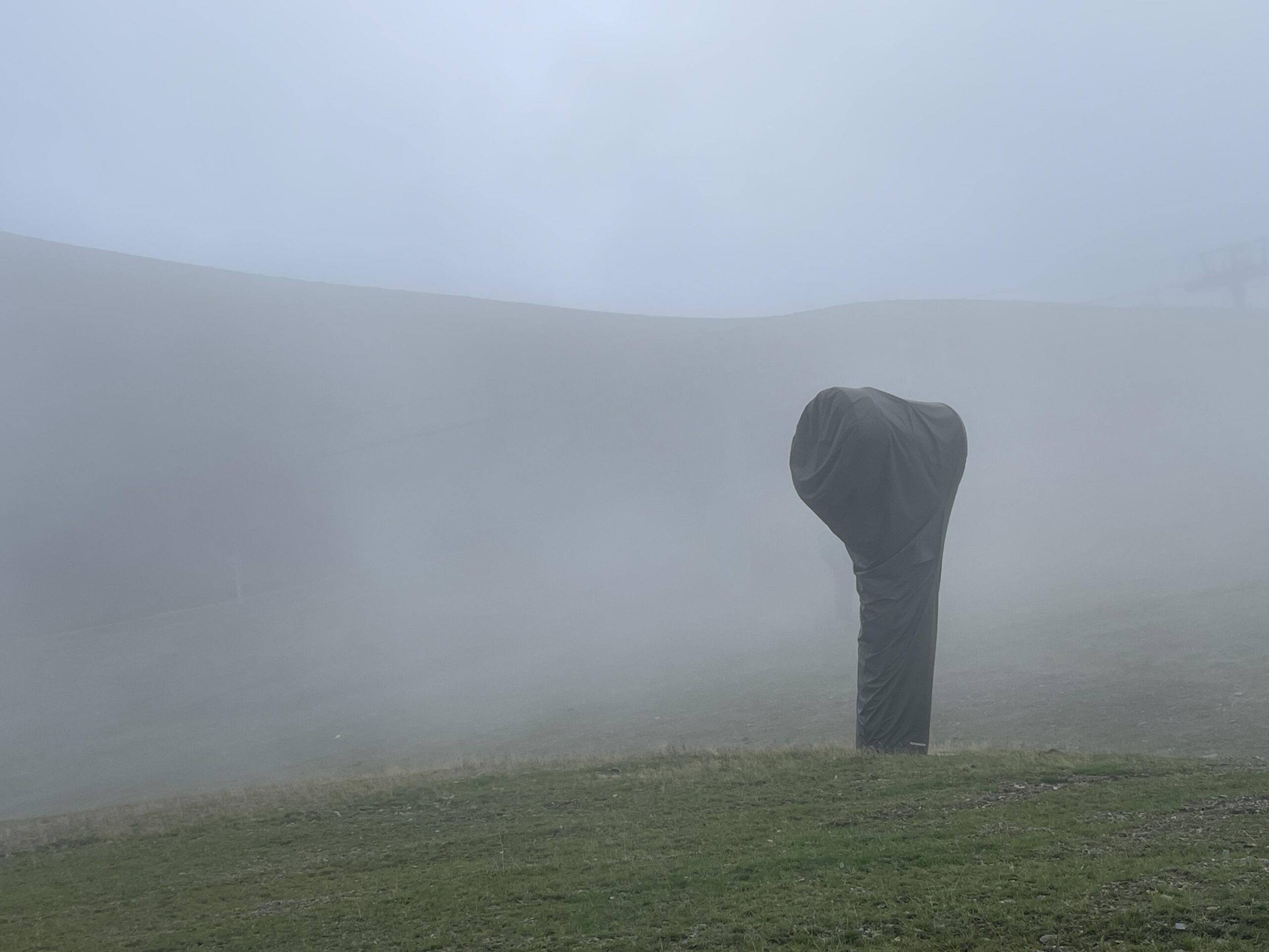 Landschaftsbild im Nebel. Im Vordergrund ist ein gras bewachsene Fläche zu sehen. sie ist leicht nach oben gekämmt so dass sie einen kleinen geschwungen Hügel bildet. Darauf steht in der rechten Seite ein großes schwarzes oder dunkelgraues Gebilde. Es besteht aus einem geraden Fuß wie eine Säue, oben verbreitert sich das Gebilde zu einem Kopf der ganz grob an die Form eines In-Ears Kopfhörers oder eines Kopfes einer elektrischen Zahnbürste erinnert. Dieser Kopf zeigt nach links. die vordere Fläche scheint rund zu sein. Das Gebilde hat kaum Struktur, bei näherem Hinsehen kann man einige Falten sehen. Dahinter ist vage im Nebel die Linie eines Hügelskamms zu sehen. Am oberen Rand in einem graublau vermutlich der Himmel