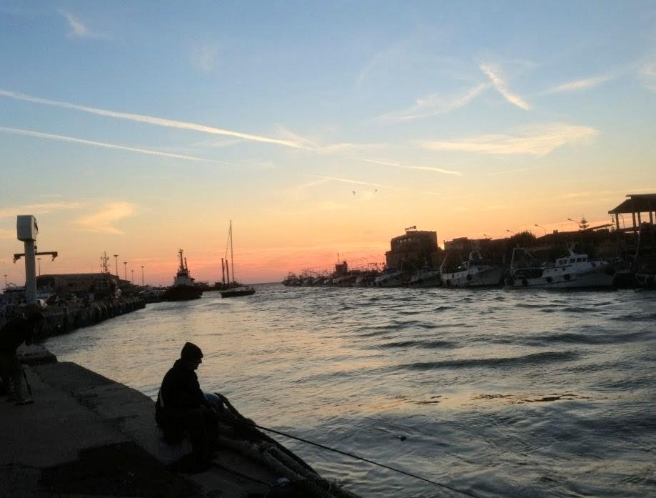 Silhouette eines Anglers auf einer Hafenmauer bei Sonnenuntergang in Fiumicino bei Rom. Das leicht bewegte Wasser führt zwischen beidseitig festgemachten Fischerbooten hinaus zum offenen Meer. Der Himmel leuchtet in sanften Orange-, Rosa- und Blautönen, durchzogen von feinen Wolken und Kondensstreifen. Im Gegenlicht zeichnen sich Gebäude, Masten und Kräne als dunkle Konturen ab.