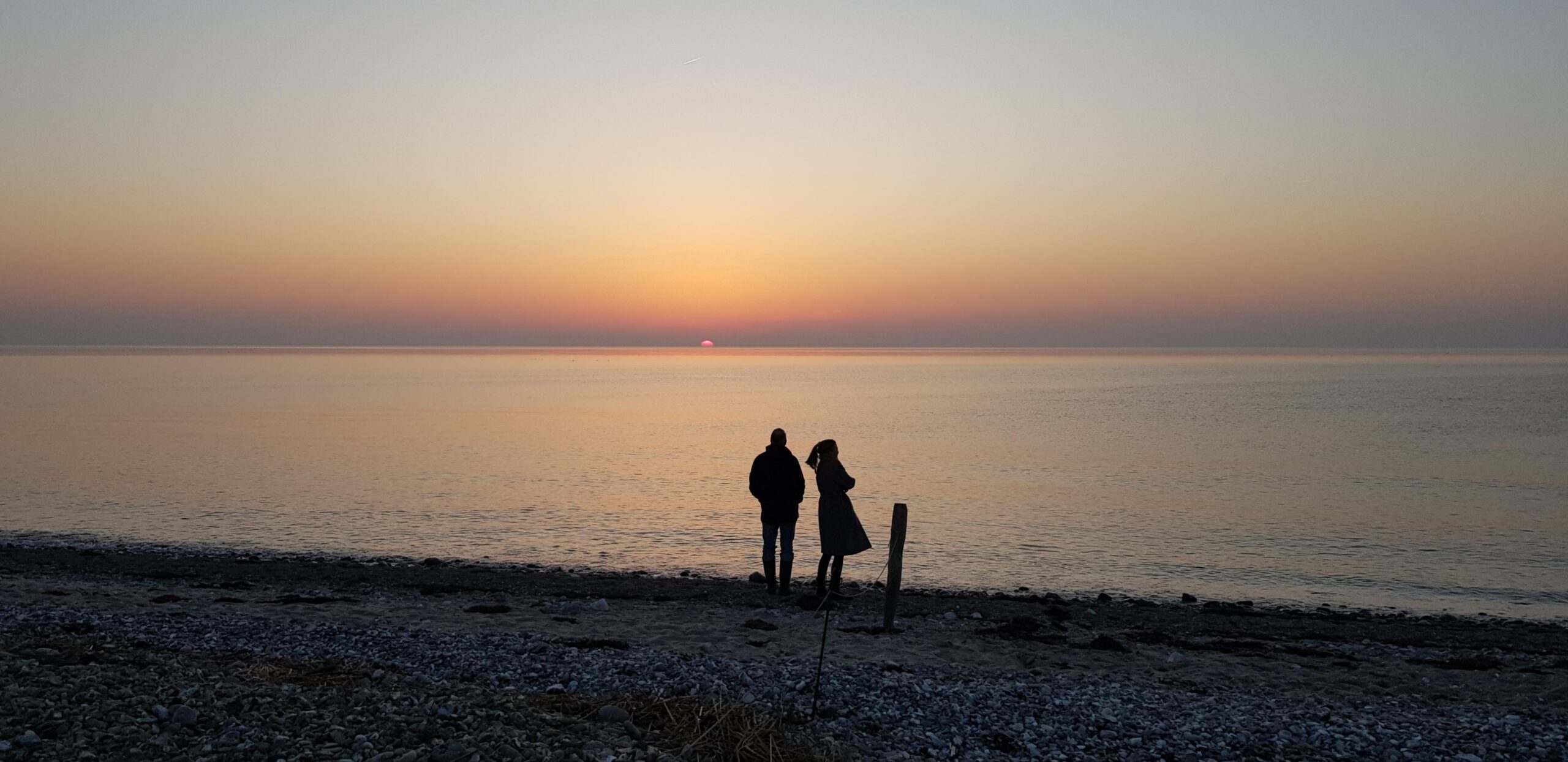 Zwei Personen stehen als schwarze Silhouetten an einem steinigen Ufer. Die linke Person hat eine eher männliche Statur, steht mit dem Rücken zum Fotografen, hat die Hände in den Hosentaschen und schaut hinaus aufs Wasser zur untergehenden Sonne, die ein blassrosa Licht über die ansonsten eher graue Szene schickt. Rechts daneben steht mit etwas Abstand eine Person mit weiblicher Statur. Durch eine Drehbewegung nach rechts schwingen Pferdeschwanz und Mantel. Die Arme sind vor dem Körper verschränkt. Der Himmel ist klar mit einem nebligen Streifen über dem Horizont. Das Wasser ist fast unbewegt. Das Ufer liegt schon im Dunkeln.
