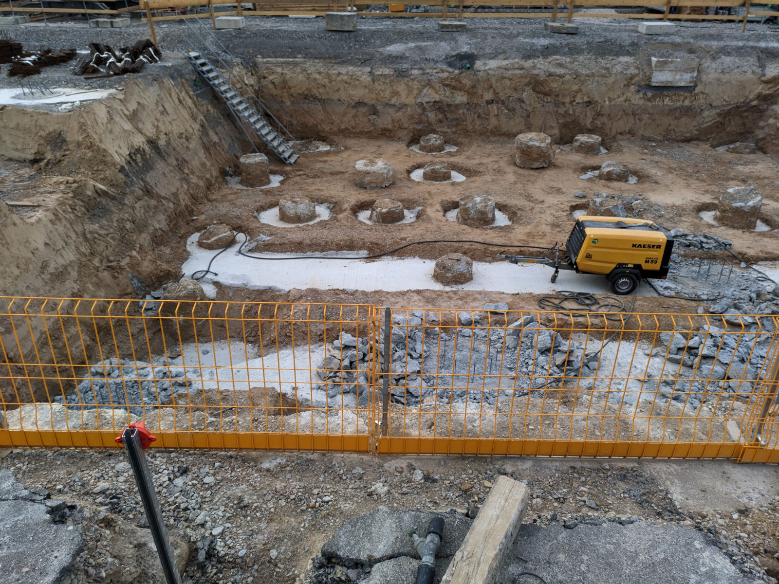 Blick über eine gelbe Abzäunung hinweg in eine Baugrube. In der linken hinteren Ecke führt eine Leiter hinab, neben dem oberen Ende der Leiter liegt rostendes Metall. In der großen Grube steht etwas verloren eine gelbe kleine Baumaschine in Anhängerform. Vom Boden der Grube ragen erdfarbene, teilweise abgebrochen wirkende Säulenstummel mit groben Oberflächen nach oben. Sie scheinen ringförmig von Beton umgeben zu sein.