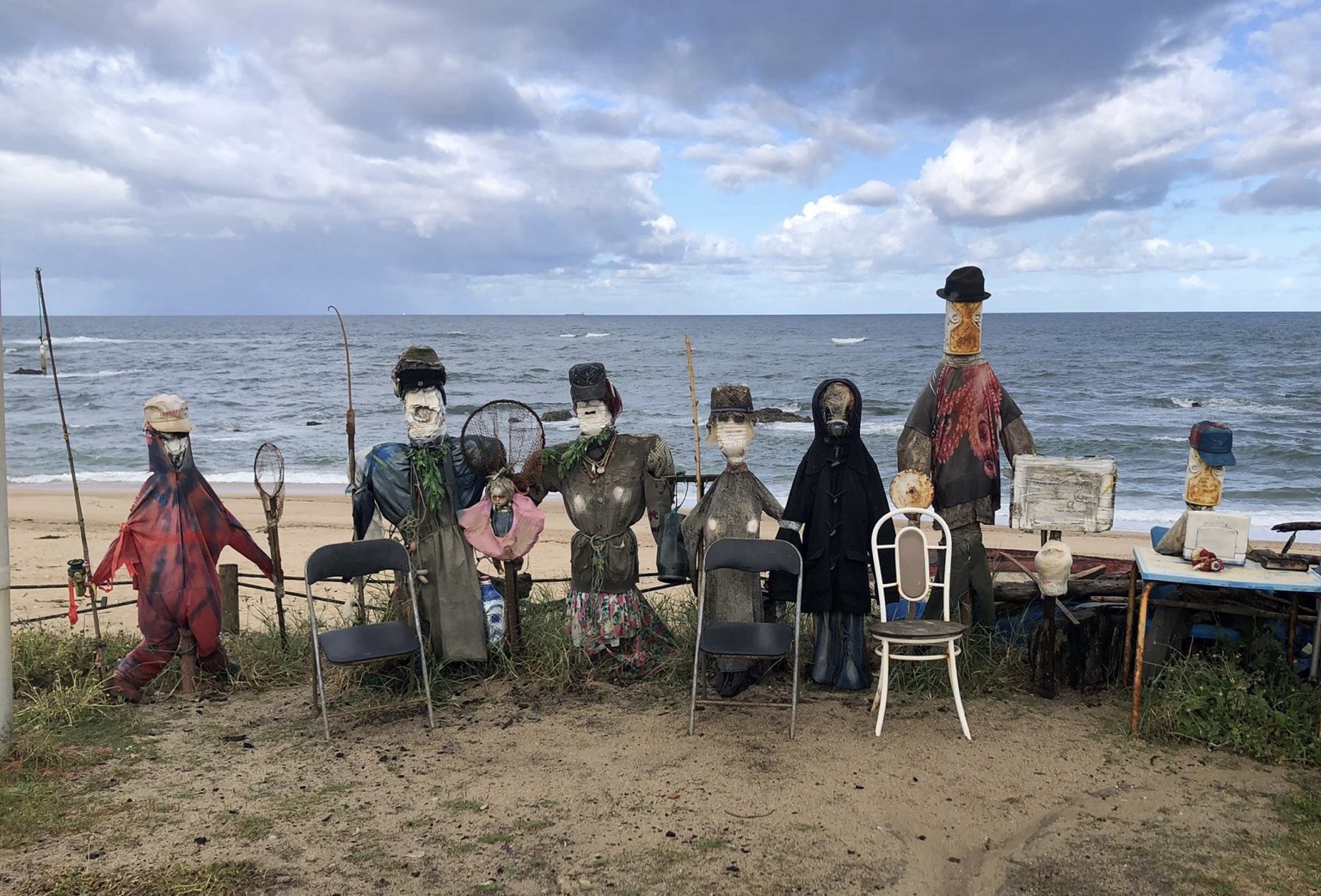 Eine Gruppe aus sieben fantasievoll gestalteten Figuren aus Stoff, Holz und anderen Materialien steht am sandigen Strand vor dem Meer. Die Figuren tragen unterschiedliche Kleidung und Hüte; einige haben verhüllte Gesichter. Zwischen ihnen stehen Stühle, Stangen und weitere Gegenstände. Im Hintergrund sind das wellige Meer und der bewölkte Himmel zu sehen.