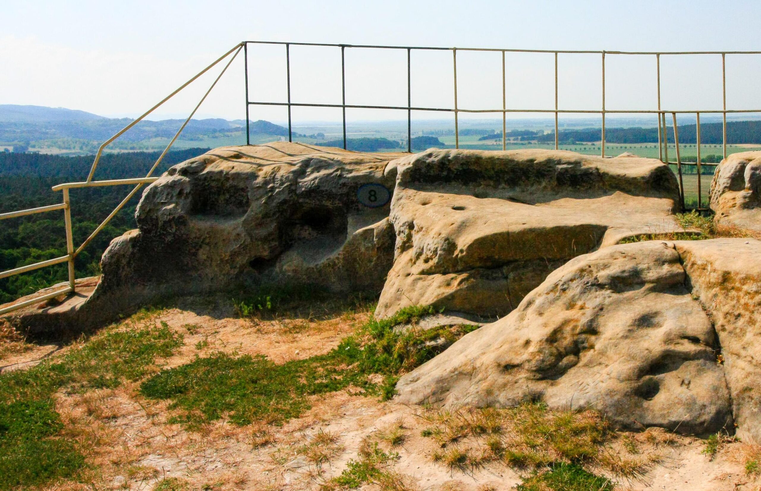 Ein nackter Fels dient als Aussichtspunkt über eine eher flache Landschaft mit Wäldern und Wiesen und der Andeutung eines Gebirges zur Linken. Ein hohes Gitter sorgt für Sicherheit.